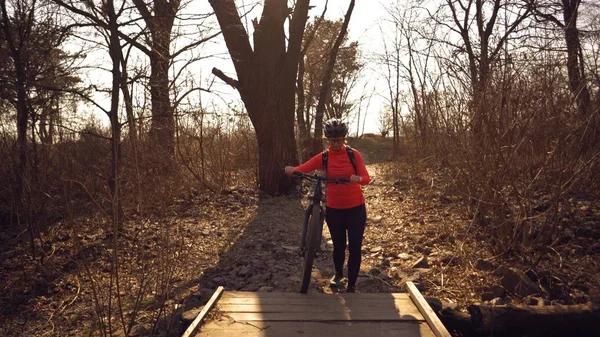 Atleta ciclista mujer caucásica en ropa deportiva y cruces de casco a pie, lleva una bicicleta de montaña en sus manos a través de un estrecho puente de madera rural a través de un pequeño río en el bosque — Foto de Stock