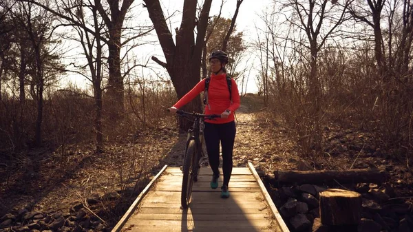 Atleet wielrenner Kaukasische vrouw in sportkleding en helm kruist te voet, leidt een mountainbike in haar handen over een landelijke smalle houten brug over een kleine rivier in het bos — Stockfoto