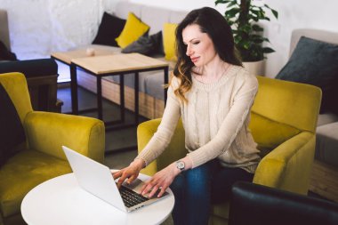 Orta yaşlı, uzun saçlı, beyaz bir kadın. İç mekanda dizüstü bilgisayar kullanarak günlük giysiler. Sandalye ahşap masanın üzerinde bir kafe restoranı. Serbest meslek yazarı, blogcu ve yazar. Klavye yazan eller