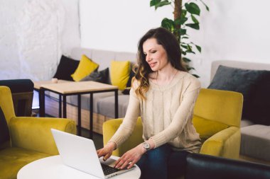 Orta yaşlı, uzun saçlı, beyaz bir kadın. İç mekanda dizüstü bilgisayar kullanarak günlük giysiler. Sandalye ahşap masanın üzerinde bir kafe restoranı. Serbest meslek yazarı, blogcu ve yazar. Klavye yazan eller