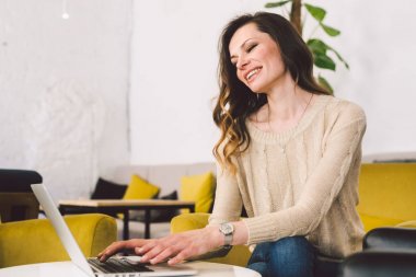 Orta yaşlı, uzun saçlı, beyaz bir kadın. İç mekanda dizüstü bilgisayar kullanarak günlük giysiler. Sandalye ahşap masanın üzerinde bir kafe restoranı. Serbest meslek yazarı, blogcu ve yazar. Klavye yazan eller