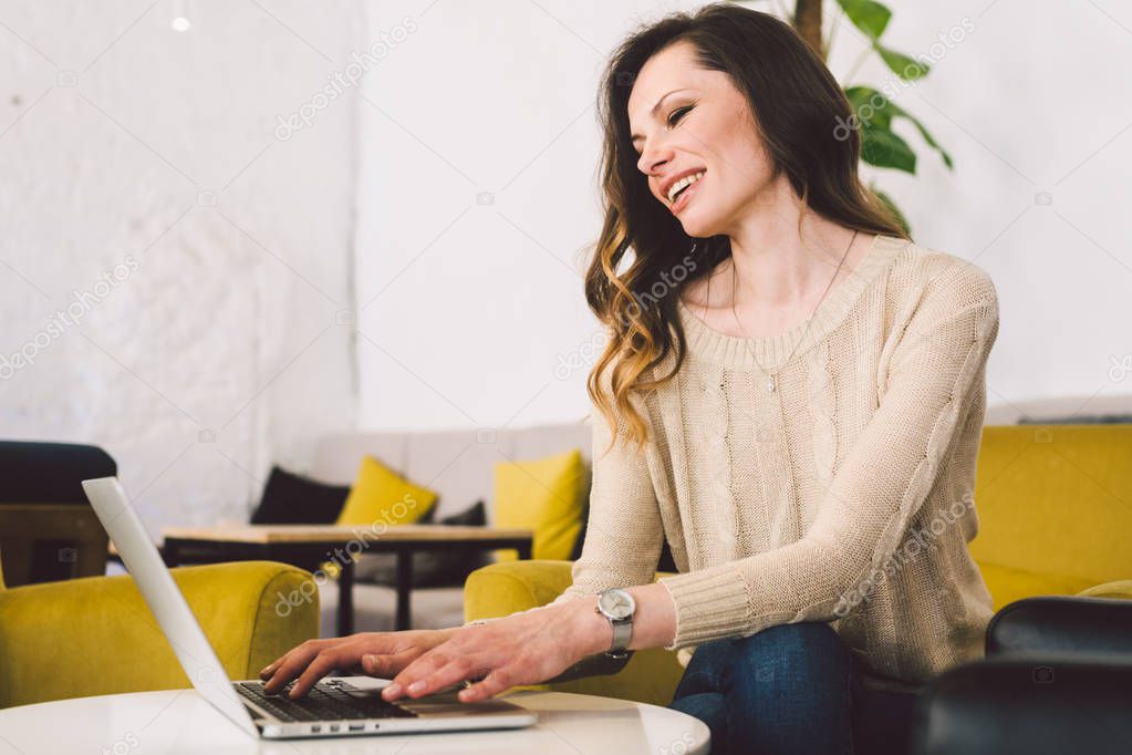 Young middle aged Caucasian woman with long hair. Casual clothes using laptop computer indoors. Cafe restaurant on chair wooden table. Profession freelancer, blogger and writer. Hands typing keyboard
