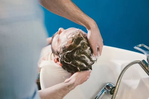 Tema da barbearia. Cabeleireiro para lavar o cabelo em uma barbearia. Mãos cabeleireiro masculino close-up lava o cabelo com espuma e shampoo homem caucasiano com barba — Fotografia de Stock