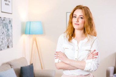 Genç beyaz kız ofiste duruyor ve kollarını göğsüne kavuşturuyordu. Çekici genç kız çalışma odasında, aydınlık ofiste çalışıyor. Kızın sarı kırmızı saçları ve beyaz gömleği var.