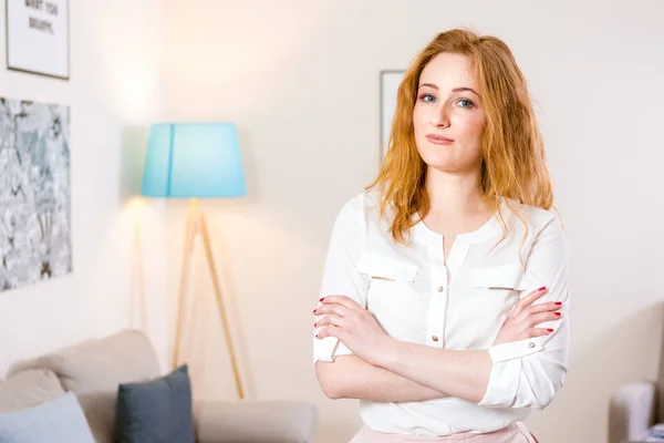 Young Caucasian girl standing in the office and folded her arms across her chest. Charming young girl works inside in bright office, coworking room. The girl has blond red hair and white shirt — ストック写真