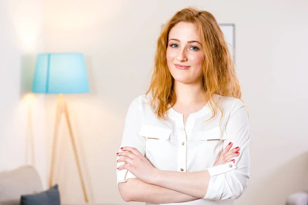 Young Caucasian girl standing in the office and folded her arms across her chest. Charming young girl works inside in bright office, coworking room. The girl has blond red hair and white shirt — ストック写真