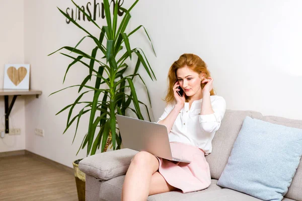 Mujer feliz sentada en el sofá con el ordenador portátil y hablando por teléfono en casa. Joven empresaria exitosa que trabaja desde casa mientras habla por teléfono. Estudiante universitario estudiando en el portátil y usando el teléfono — Foto de Stock