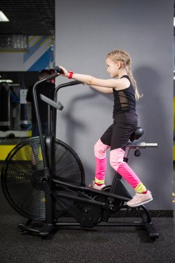 Spor salonunda egzersiz bisikleti kullanan küçük çekici beyaz çocuk. Fitness. Crossfit spor salonunda kardiyo egzersiz için bir hava bisikleti kullanan küçük bir atlet. Bisiklet makinesinde oturan spor kız