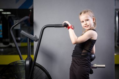 Spor salonunda egzersiz bisikleti kullanan küçük çekici beyaz çocuk. Fitness. Crossfit spor salonunda kardiyo egzersiz için bir hava bisikleti kullanan küçük bir atlet. Bisiklet makinesinde oturan spor kız