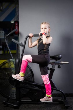 Spor salonunda egzersiz bisikleti kullanan küçük çekici beyaz çocuk. Fitness. Crossfit spor salonunda kardiyo egzersiz için bir hava bisikleti kullanan küçük bir atlet. Bisiklet makinesinde oturan spor kız