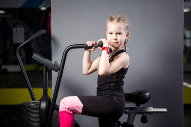 Spor salonunda egzersiz bisikleti kullanan küçük çekici beyaz çocuk. Fitness. Crossfit spor salonunda kardiyo egzersiz için bir hava bisikleti kullanan küçük bir atlet. Bisiklet makinesinde oturan spor kız