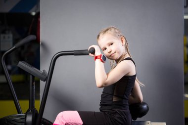 Spor salonunda egzersiz bisikleti kullanan küçük çekici beyaz çocuk. Fitness. Crossfit spor salonunda kardiyo egzersiz için bir hava bisikleti kullanan küçük bir atlet. Bisiklet makinesinde oturan spor kız