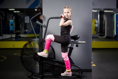 Spor salonunda egzersiz bisikleti kullanan küçük çekici beyaz çocuk. Fitness. Crossfit spor salonunda kardiyo egzersiz için bir hava bisikleti kullanan küçük bir atlet. Bisiklet makinesinde oturan spor kız