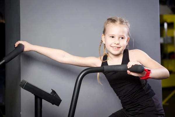 Pequena criança caucasiana atraente usando bicicleta de exercício no ginásio. Aptidão. Um pequeno atleta usando uma bicicleta de ar para um treino de cardio no ginásio crossfit. Esporte menina sentada na máquina de bicicleta — Fotografia de Stock