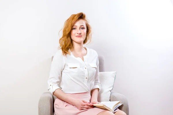 Young beautiful caucasian woman with long magnificent red hair and freckles on face. Student girl sitting on armchair and holding book in hands, a man reading a book indoors on a white background — ストック写真