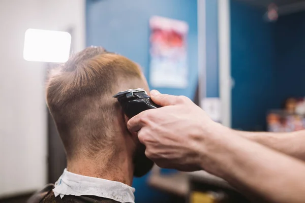 Tiro de perto de um homem a cortar o cabelo na barbearia. Cabeleireiro masculino servindo cliente, fazendo corte de cabelo usando máquina e pente — Fotografia de Stock