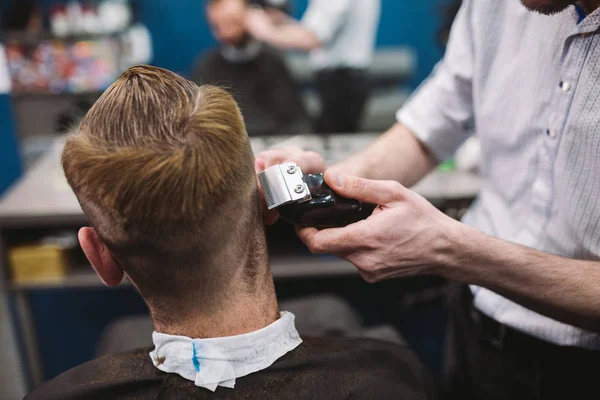 Крупним планом чоловік стає модною стрижкою в перукарні. Чоловічий перукар, що обслуговує клієнта, робить зачіску за допомогою машини та гребеня — стокове фото