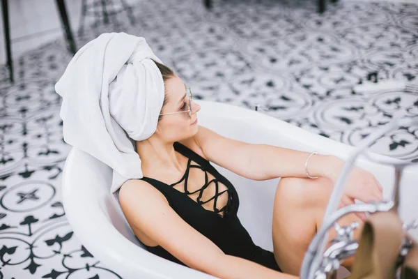 Νέα καυκάσια γυναίκα με πετσέτα στο κεφάλι και τα γυαλιά ηλίου να πάρει θεραπεία σπα σε ένα σαλόνι ομορφιάς, μέσα σε ένα εσωτερικό δωμάτιο. Χαλαρώστε στο μπάνιο με τα λευκά είδη. Η έννοια της φροντίδας του σώματος και της χαλάρωσης — Φωτογραφία Αρχείου