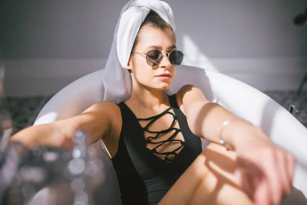 Junge kaukasische Frau mit Handtuch auf dem Kopf und Sonnenbrille, die sich in einem Schönheitssalon in einem Innenraum einer Wellnessbehandlung unterzieht. Entspannung im Badezimmer in Bettwäsche. das Konzept der Körperpflege und Entspannung — Stockfoto