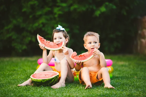 Divertente fratellino e sorella che mangiano anguria su erba verde vicino alla piscina gonfiabile in cortile a casa. Maschietto e femminuccia. I bambini mangiano frutta in giardino. Infanzia, Famiglia, Dieta sana — Foto Stock