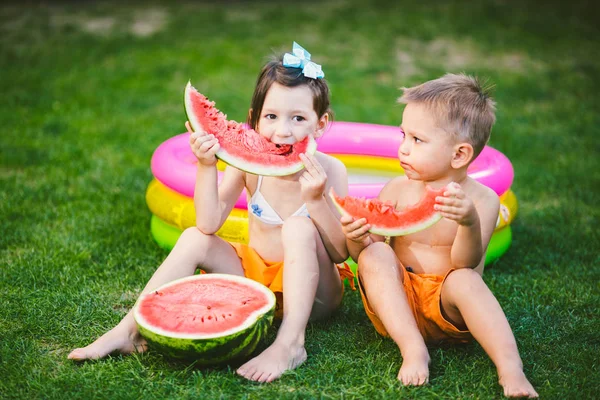 Divertente fratellino e sorella che mangiano anguria su erba verde vicino alla piscina gonfiabile in cortile a casa. Maschietto e femminuccia. I bambini mangiano frutta in giardino. Infanzia, Famiglia, Dieta sana — Foto Stock