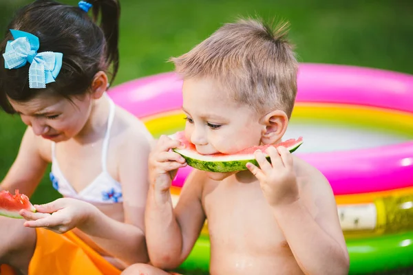 Divertente fratellino e sorella che mangiano anguria su erba verde vicino alla piscina gonfiabile in cortile a casa. Maschietto e femminuccia. I bambini mangiano frutta in giardino. Infanzia, Famiglia, Dieta sana — Foto Stock