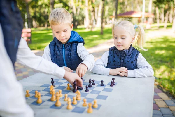 O tema crianças aprendizagem, desenvolvimento lógico, mente e matemática, erros de cálculo avança. grande família irmãos e irmã caucasiano meninos e menina jogando xadrez parque brilhante sol tempo outono — Fotografia de Stock