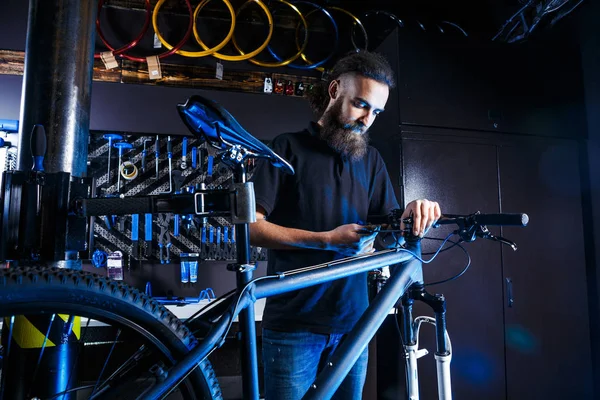 Tema venta y reparación de bicicletas. Joven y elegante con barba y pelo largo, un hombre caucásico utiliza una herramienta para montar y reparar una bicicleta en una tienda. Propietario del negocio en el trabajo —  Fotos de Stock