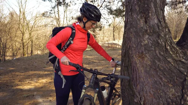 Кавказская спортсменка-велосипедистка на внедорожнике возле дерева. Спортсменка в спортивной одежде, шлеме и черном рюкзаке останавливается на горном велосипеде — стоковое фото