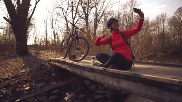 Vrolijke blanke vrouw fietser maakt een foto van zichzelf een selfie aan de telefoon tijdens het zitten op de brug over een rivier in het bos bij zonnig weer — Stockvideo