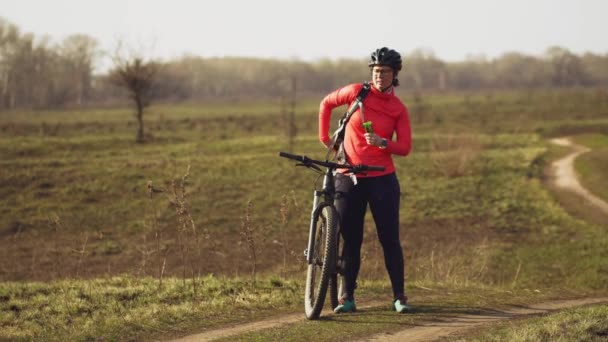 Mulher branca atlética come passeio de bar de proteína em mountain bike na natureza. Jovem atleta esportiva no capacete descansando enquanto morde a barra nutricional. Fitness mulher comer energia lanche ao ar livre — Vídeo de Stock