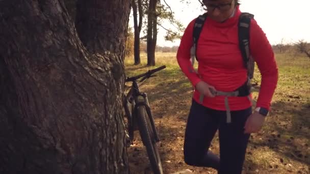 Caucasian sport cyclist woman sport off road bike park near tree. woman athlete in sport clothes and helmet and black backpack stop to sit back in nature and use mobile phone technology in her hand — Stock Video