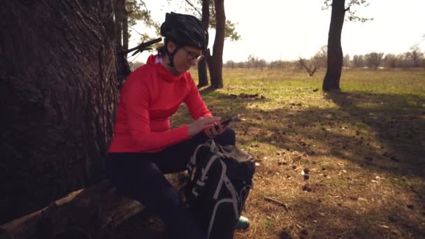 Caucasian sport cyclist woman sport off road bike park near tree. woman athlete in sport clothes and helmet and black backpack stop to sit back in nature and use mobile phone technology in her hand — Stock Video