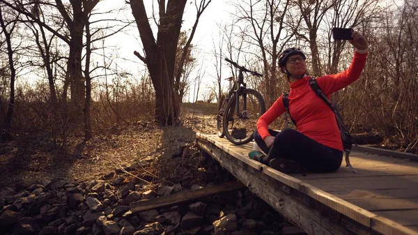 Vrolijke blanke vrouw fietser maakt een foto van zichzelf een selfie aan de telefoon tijdens het zitten op de brug over een rivier in het bos bij zonnig weer — Stockfoto