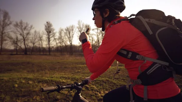 Спортивная белая женщина ест протеиновый бар на горном велосипеде на природе. Молодая спортсменка в шлеме отдыхает, кусая диетический бар. Фитнес-женщина ест энергетические закуски на открытом воздухе — стоковое фото