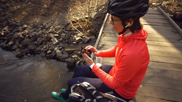 Mujer ciclista deportiva caucásica escuchando música en auriculares blancos inalámbricos sentados en un puente de madera sobre un pequeño río en la naturaleza en ropa deportiva y un casco cerca de una mochila negra — Foto de Stock