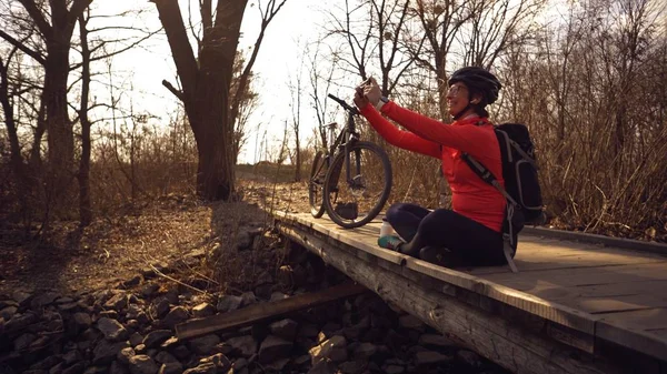 Feliz mujer caucásica ciclista hace una foto de sí misma una selfie en el teléfono mientras está sentada en el puente sobre un río en el bosque en tiempo soleado — Foto de Stock