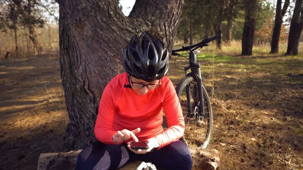 Kaukaski Sport rowerzysta kobieta sportu Off Road Bike Park w pobliżu drzewa. Kobieta sportowiec w odzieży sportowej i kask i czarny plecak przystanek siedzieć z powrotem w przyrodzie i korzystać z technologii telefonu komórkowego w ręku — Zdjęcie stockowe