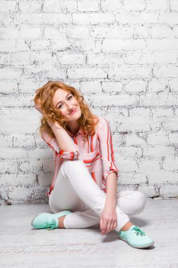 Young beautiful caucasian woman student is sitting on the floor near a white brick wall. Girl with red long curly hair with freckles on her face in bright summer clothes