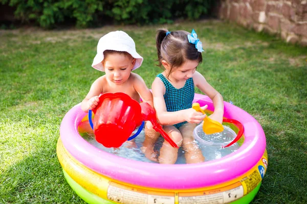 Thema Kindersommerspiele im Hof. Kaukasisches Geschwisterpaar spielt Plastik-Spielzeug-Eimer im Wasser, aufblasbares rundes Kinderbecken. Sommer ist heiß, Ruhe in Badeanzügen — Stockfoto