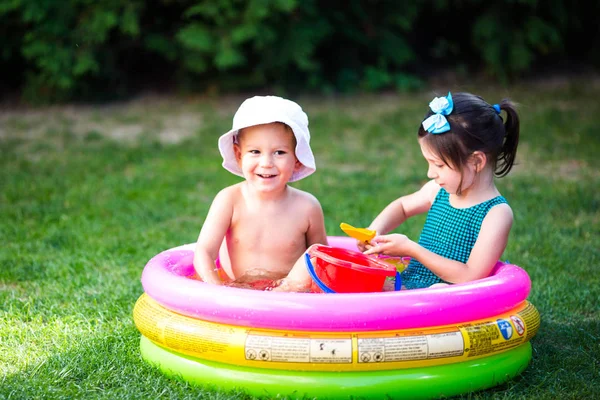 庭で子供の頃の夏のゲームを対象。白人の兄弟姉妹は、水の中に座ってプラスチックのおもちゃのバケツを再生し、膨脹可能な丸い子供のプール。夏は暑く、水着で休む — ストック写真