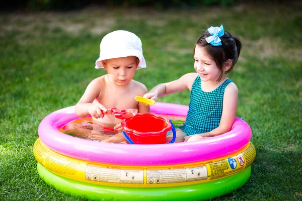 Bahçede Konu çocukluk yaz oyunları. Beyaz abi ve kız kardeşi plastik oyuncak kova suda oturan oynarken, şişme yuvarlak çocuk havuzu. Yaz sıcak, mayolarla dinlenin — Stok fotoğraf