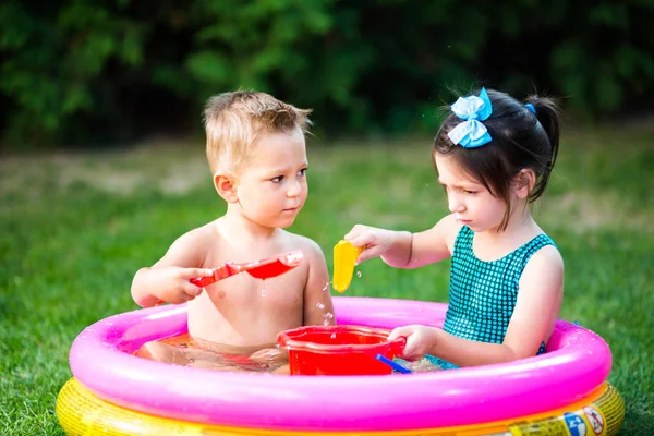 Детские летние игры во дворе. Кавказский брат и сестра играют в пластиковые игрушки ведро сидя в воде, надувной круглый детский бассейн. Лето жарко, отдых в купальниках — стоковое фото
