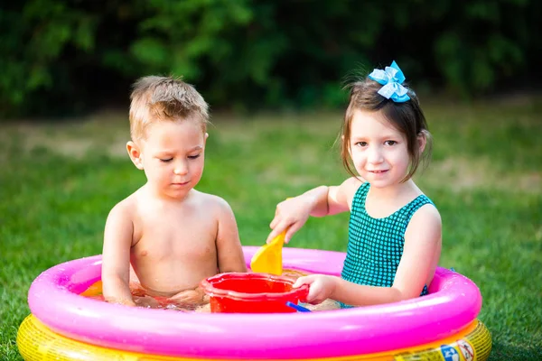 Thema Kindersommerspiele im Hof. Kaukasisches Geschwisterpaar spielt Plastik-Spielzeug-Eimer im Wasser, aufblasbares rundes Kinderbecken. Sommer ist heiß, Ruhe in Badeanzügen — Stockfoto