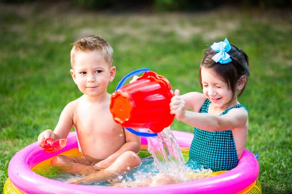 Subject childhood summer games in the yard. Caucasian brother and sister playing plastic toys bucket sitting in the water, inflatable round children\'s pool. Summer is hot, rest in swimsuits