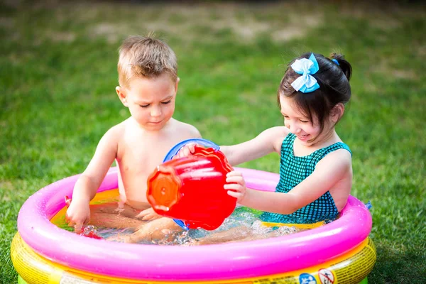 Asunto juegos de verano de la infancia en el patio. Hermano y hermana caucásicos jugando cubo de juguetes de plástico sentado en el agua, piscina inflable redonda para niños. El verano es caluroso, descansa en bañadores — Foto de Stock