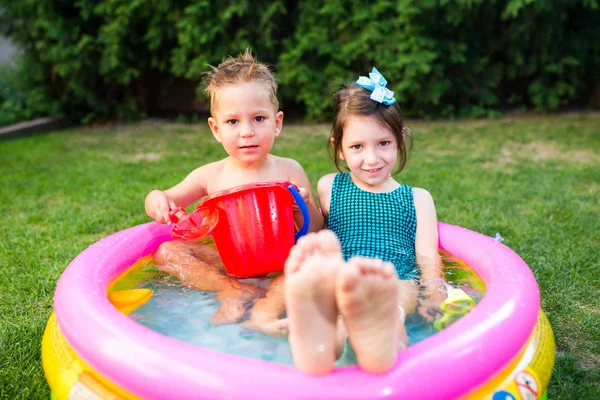庭で子供の頃の夏のゲームを対象。白人の兄弟姉妹は、水の中に座ってプラスチックのおもちゃのバケツを再生し、膨脹可能な丸い子供のプール。夏は暑く、水着で休む — ストック写真