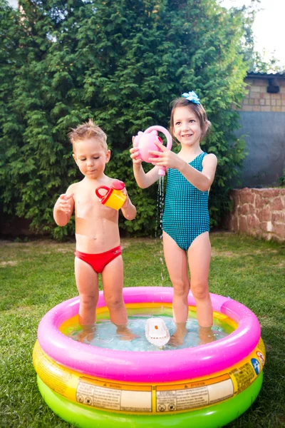 childhood summer games with water pool. Caucasian brother and sister play with plastic toys watering can pouring water splashing, inflatable round childrens bathroom. Summer hot holidays in swimsuits