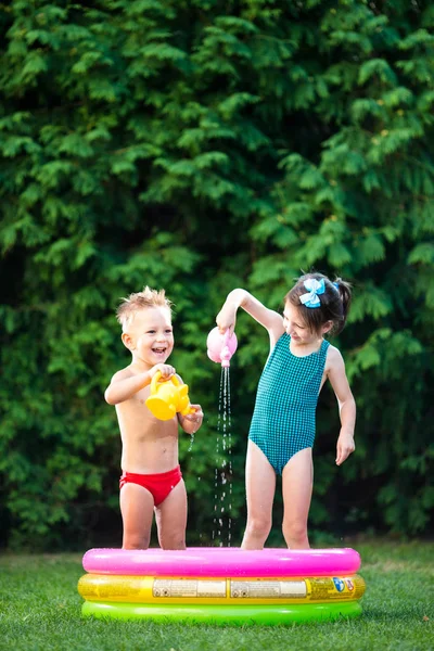 childhood summer games with water pool. Caucasian brother and sister play with plastic toys watering can pouring water splashing, inflatable round childrens bathroom. Summer hot holidays in swimsuits