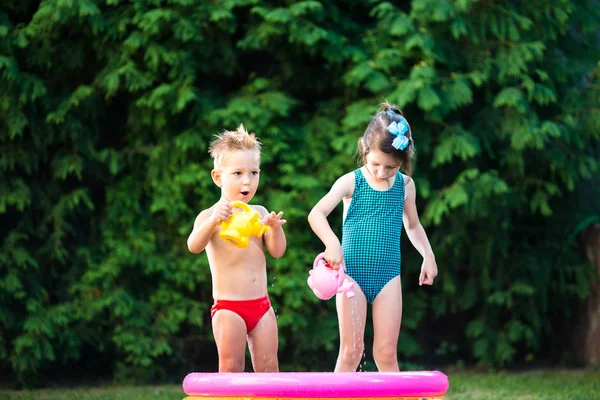 childhood summer games with water pool. Caucasian brother and sister play with plastic toys watering can pouring water splashing, inflatable round childrens bathroom. Summer hot holidays in swimsuits